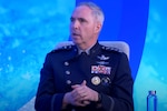 A man in a military dress uniform sits in a chair and speaks to a crowd.