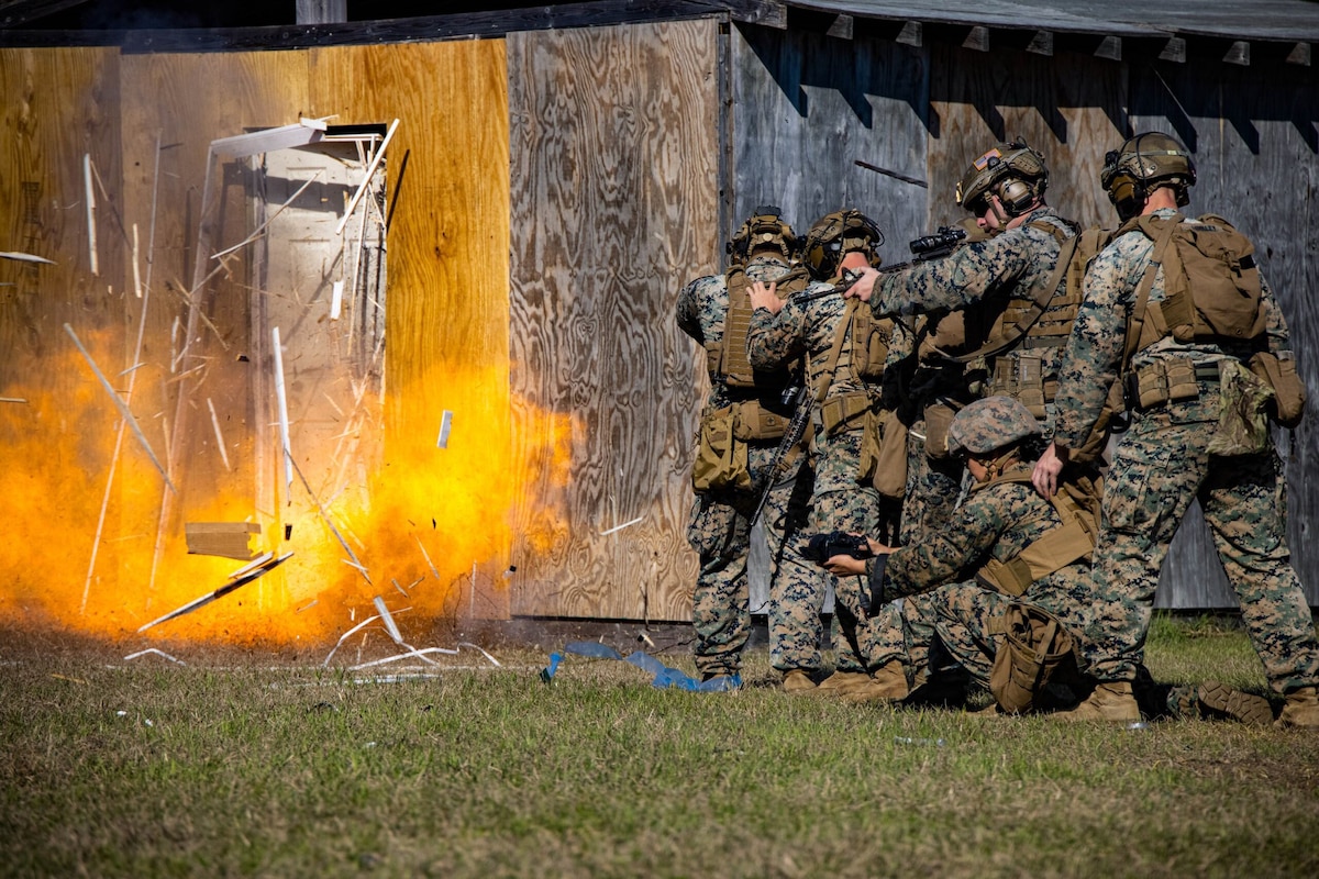 Marines in camouflage uniforms stand in formation as a door in front of them explodes.