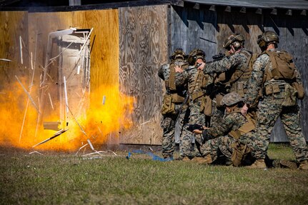 Marines in camouflage uniforms stand in formation as a door in front of them explodes.