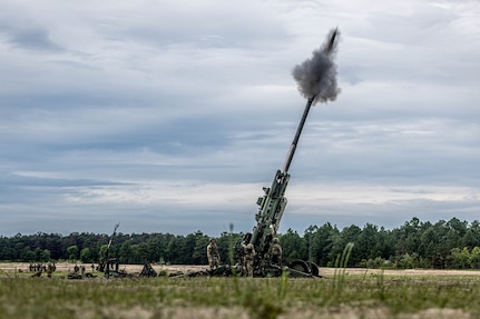 A large gun fires into the air as service members in camouflage uniforms stand around it.