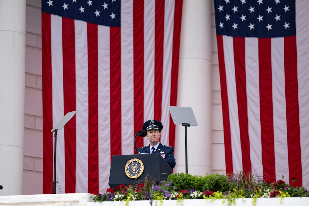The chairman speaks on a stage in front of a large hanging American flag.