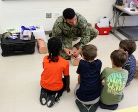 Hospital Corpsman Second Class (HM2) Ethan Johnson utilized training equipment from Tactical Combat Casualty Care (TCCC) to demonstrate wound packing and bandaging techniques at the Captain Walter Francis Duke Elementary School's Career Day. Johnson provided a brief overview of TCCC and the critical role corpsmen play in stabilizing Marines and Sailors in combat situations until they are transported for more advanced medical care. Students were given the opportunity to actively work with the TCCC equipment themselves, practicing wound care and bandaging techniques