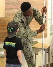 Hospital Corpsman Third Class (HM3) Rickey Samuels, an Aerospace Medical Technician, demonstrated how pilot candidates are carefully measured and assessed. Samuels highlighted the crucial role of an Aerospace Medical Technician in ensuring the health and readiness of naval aviators, creating a wave of enthusiasm among the students. Eager to gauge their own potential for a career in the cockpit, this student asks about measuring their height and arm length to compare their measurements to Navy pilot standards.
