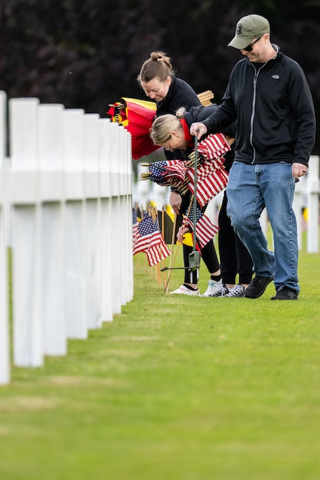 Three people stamp holes in the ground and place American and Belgian flags in front of a row of limestone cross headstones.