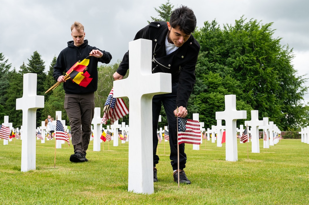 A man places an American flag in front of a limestone cross headstone while another man to frame's left pulls a Belgian flag from a bundle in his arms to put in front of a similar headstone