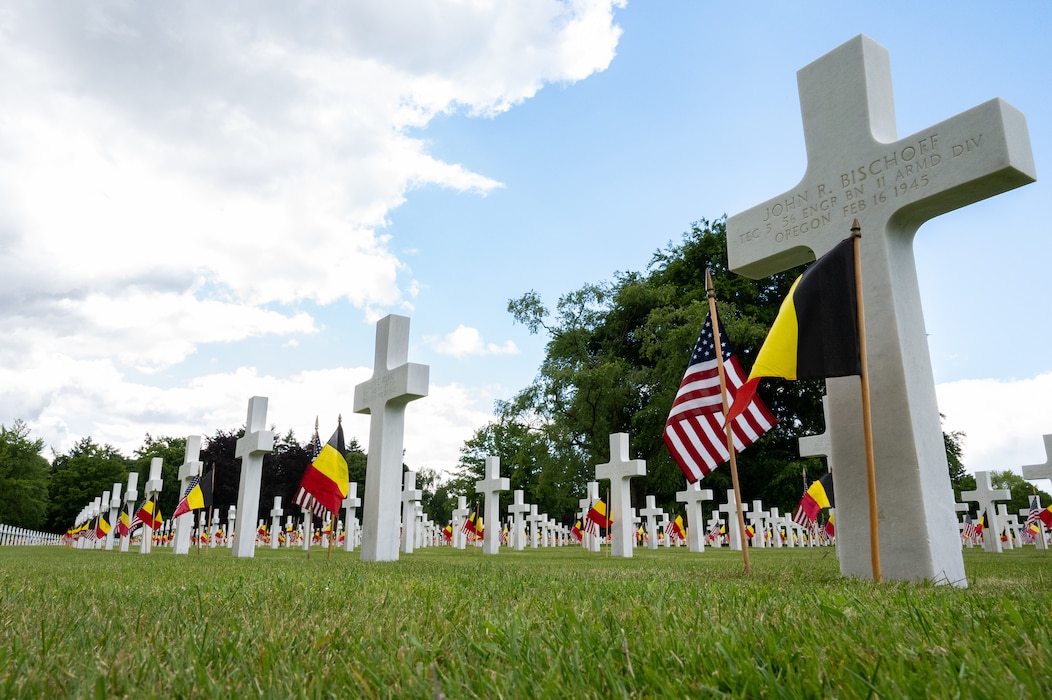 Rows of limestone cross and Star-of-David headstones with American and Belgian flags planted in front of each of grave.