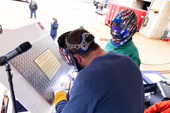 The sponsor of DDG 132, Madison Ann Zolper, who is the great-granddaughter of Quentin Walsh, strikes welding sparks into a keel plate inscribed with her initials. At left is BIW welder Eric Goldsmith who assisted the sponsor.