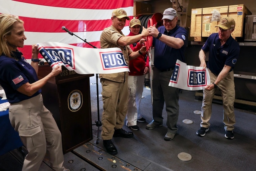 Cmdr. Raymond Miller IV, center left, commanding officer of Arleigh Burke-class guided-missile destroyer USS Bainbridge (DDG 96), and Mr. Christopher Plamp, Chief Operating Officer, United Service Organizations, cut a ribbon in the hangar bay in honor of the ship’s new USO unstaffed entertainment center, May 22, 2025. Bainbridge is a part of Carrier Strike Group 12, which provides combatant commanders and America’s civilian leaders multi-domain power to deter adversaries, underpin American security and economic prosperity, and project global power. (U.S. Navy photo by Mass Communication Specialist Seaman Cesar Licona)