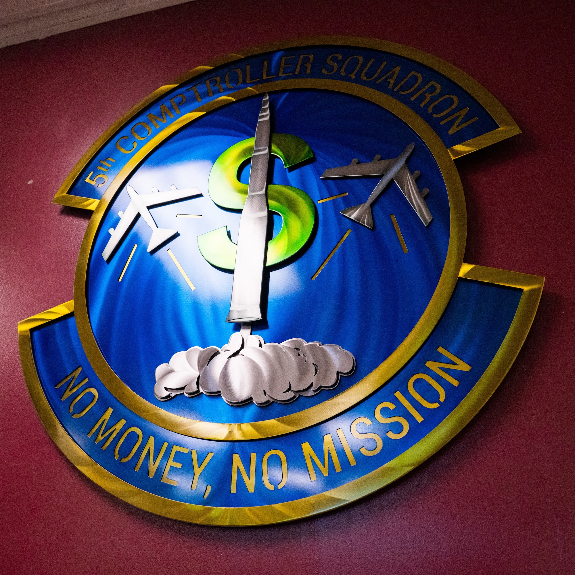 The symbol of the 5th Comptroller Squadron hanging on display. It is a circular symbol featuring a large dollar sign ($) with aircraft flying around it. The symbol has the motto, "No Money, No Mission" written below it.