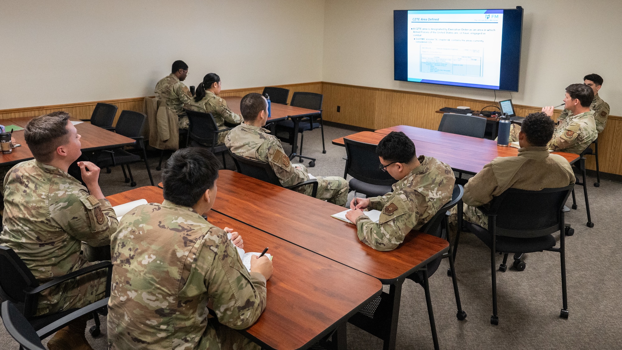 A classroom of Airmen listen to a training lecture alongside projected slides.