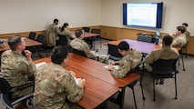 A classroom of Airmen listen to a training lecture alongside projected slides.