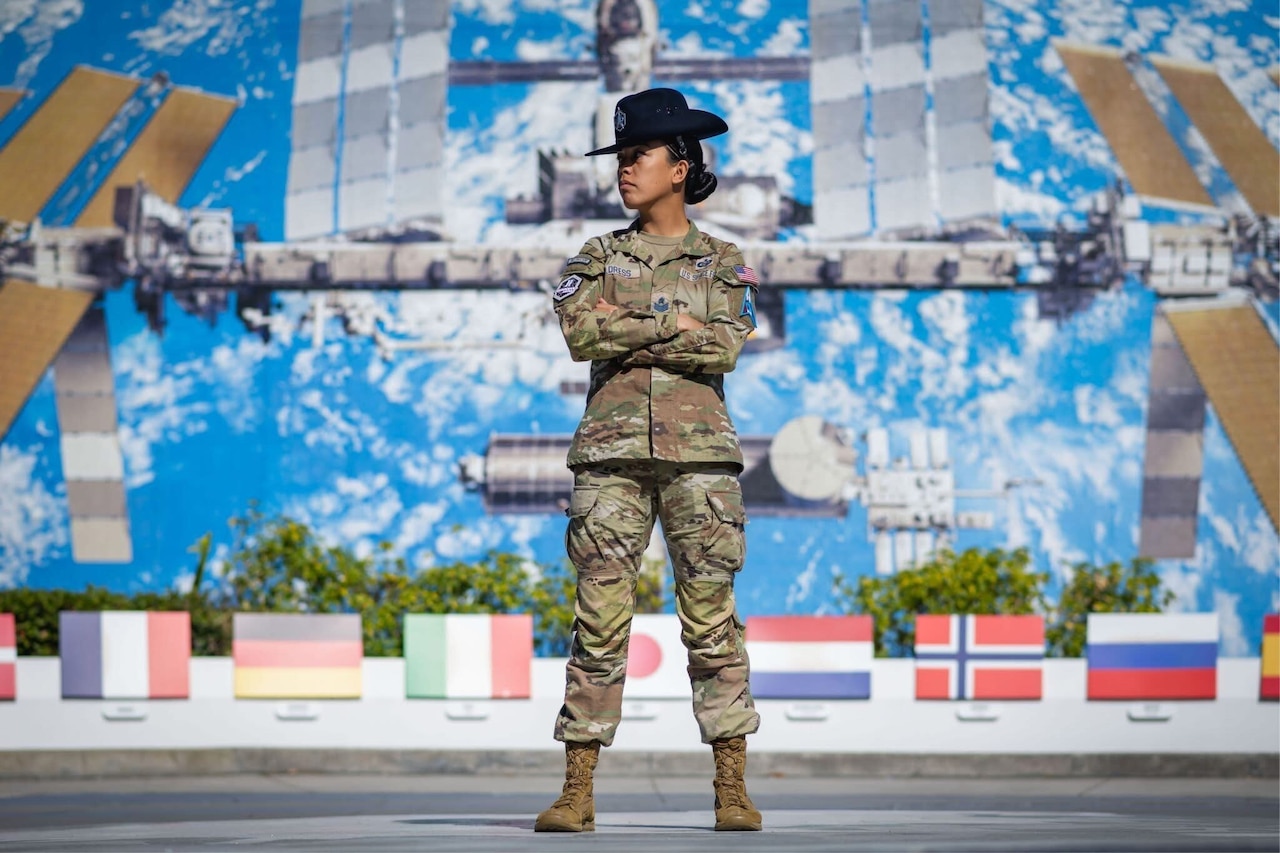 A woman in uniform stands with her arms crossed over her chest in front of a mural of a satellite.