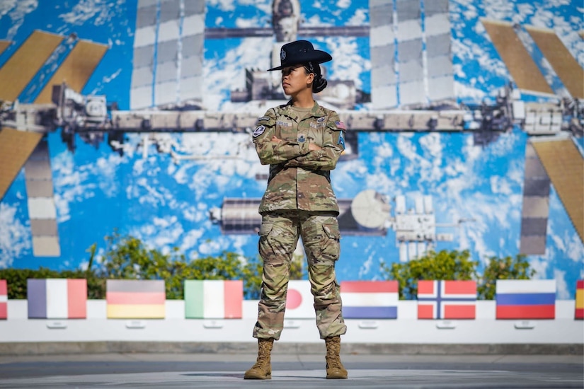 A woman in uniform stands with her arms crossed over her chest in front of a mural of a satellite.