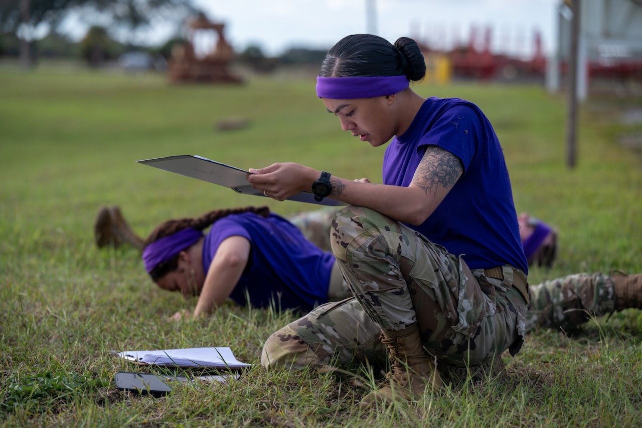 A kneeling woman holds a clipboard and looks at papers in the grass. Two others behind her are doing pushups.
