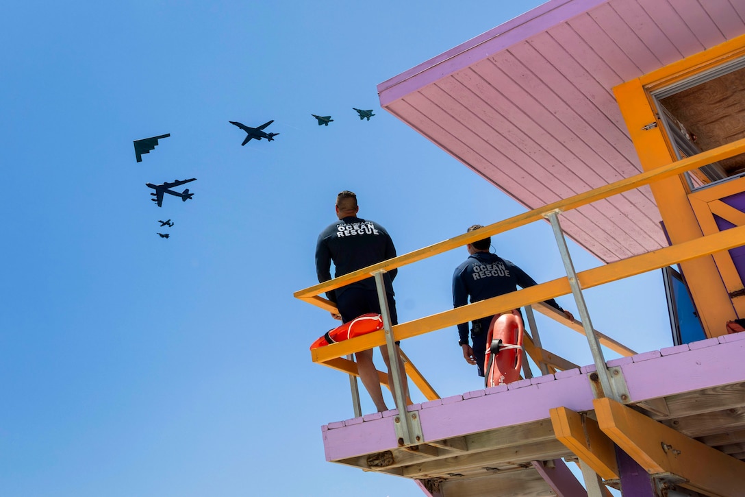 Two lifeguards stand on a purple and yellow beach station as seven aircraft fly in a blue sky.