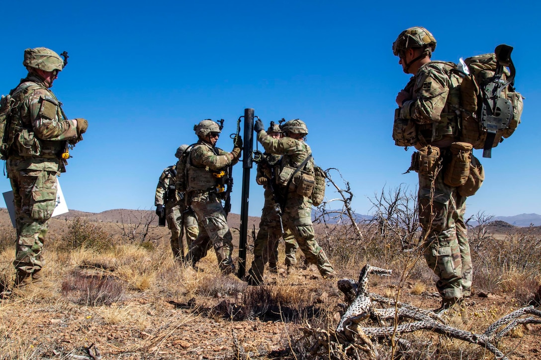 Three soldiers install a sign post in a desert-like area as three others look on with mountains and a blue sky in the background.