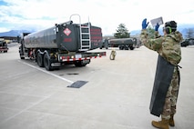 an airman with his back towards the camera stands behind a fuel truck directing it to move backwards