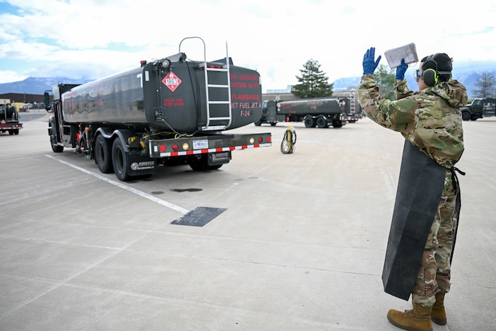 an airman with his back towards the camera stands behind a fuel truck directing it to move backwards