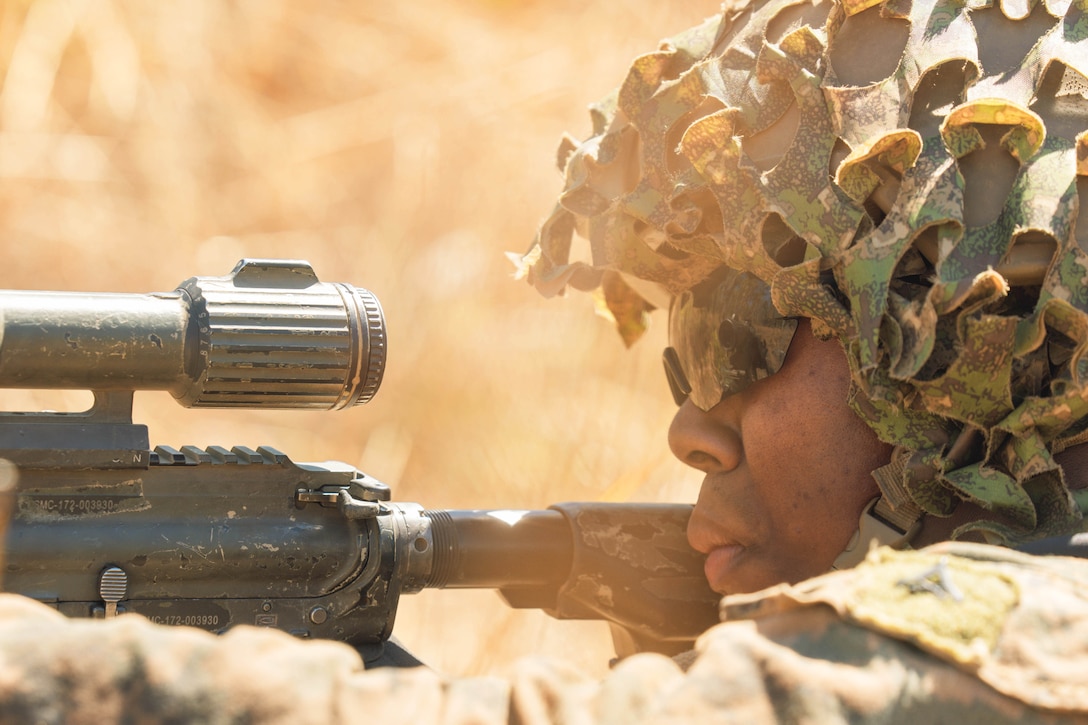 A close-up of a Marine wearing camouflage lays on the ground looking through the scope of a weapon surrounded by clouds of dust.