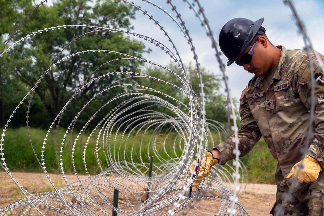 A guardsman wearing a construction helmet uses a tool to install concertina wire to a post.
