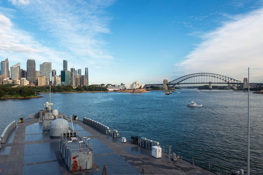 A ship's deck is seen in the foreground as it transits a body of water with a city skyline and bridge in the background under a blue, cloudy sky.