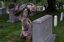 An Army Soldier dressed in combat uniform with gloves is bending over and placing a small US flag in front of a large, rectangular-shaped tombstone
