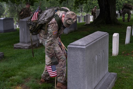 An Army Soldier dressed in combat uniform with gloves is bending over and placing a small US flag in front of a large, rectangular-shaped tombstone