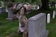 An Army Soldier dressed in combat uniform with gloves is bending over and placing a small US flag in front of a large, rectangular-shaped tombstone