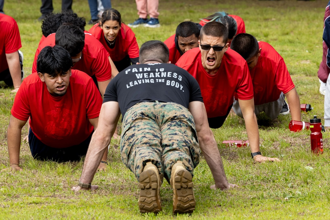 A Marine completes push-ups while facing people in red shirts on grass.