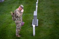 An Army soldier dressed in combat uniform is saluting towards a tombstone that has a small US flag placed in front of it.