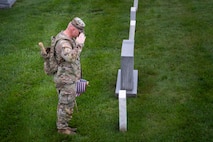 An Army soldier dressed in combat uniform is saluting towards a tombstone that has a small US flag placed in front of it.