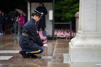 An Army Soldier dressed in dark blue ceremonial uniform with raincoat is carrying small US flags while placing one in the ground at the Tomb of the Unknown Soldier.