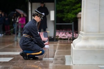 An Army Soldier dressed in dark blue ceremonial uniform with raincoat is carrying small US flags while placing one in the ground at the Tomb of the Unknown Soldier.