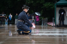 An Army Solider dressed in ceremonial dress uniform with dark blue raincoat is placing a small US flag at one of the marble crypts at the Tomb of the Unknown Soldier.