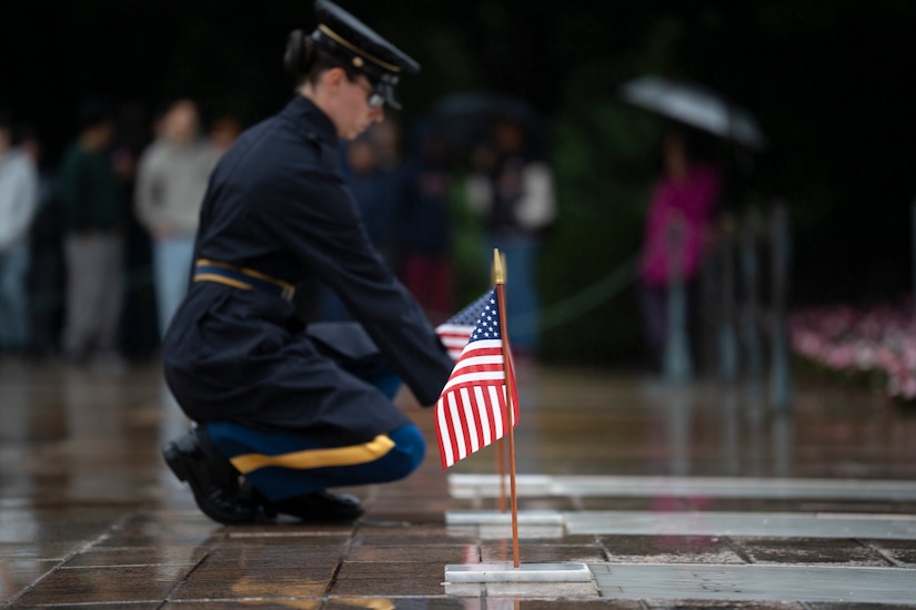 A U.S. Army Soldier wearing a dark blue ceremonial uniform with raincoat is kneeling over at the Tomb of the Unknown Soldier and placing a small U.S. flag at one of the three crypts.