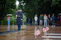 A U.S. Army Soldier wearing a ceremonial uniform with raincoat is standing at attention on a black mat surrounded by wet pavement while maintaining the watch at the Tomb of the Unknown Soldier, which has three small U.S. flags placed over the three crypts.