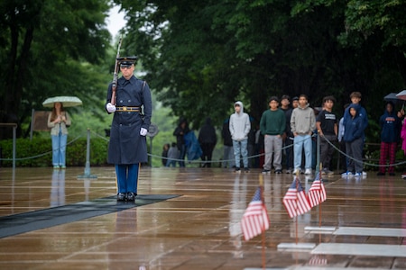 A U.S. Army Soldier wearing a ceremonial uniform with raincoat is standing at attention on a black mat surrounded by wet pavement while maintaining the watch at the Tomb of the Unknown Soldier, which has three small U.S. flags placed over the three crypts.