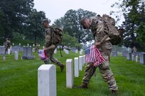 A U.S. Army Soldier wearing combat uniform is putting his foot next to a tombstone to measure the distance of where he will put a small U.S. flag. He has dozens of small U.S. flags in his hand and is walking next to another Soldier through a cemetery.