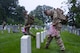 A U.S. Army Soldier wearing combat uniform is putting his foot next to a tombstone to measure the distance of where he will put a small U.S. flag. He has dozens of small U.S. flags in his hand and is walking next to another Soldier through a cemetery.