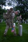 A U.S. Army Soldier wearing combat uniform is placing a small U.S. flag into the ground next to a tombstone.