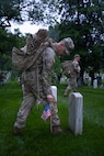 A U.S. Army Soldier wearing combat uniform is placing a small U.S. flag into the ground next to a tombstone.