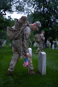 A U.S. Army Soldier wearing combat uniform is placing a small U.S. flag into the ground next to a tombstone.