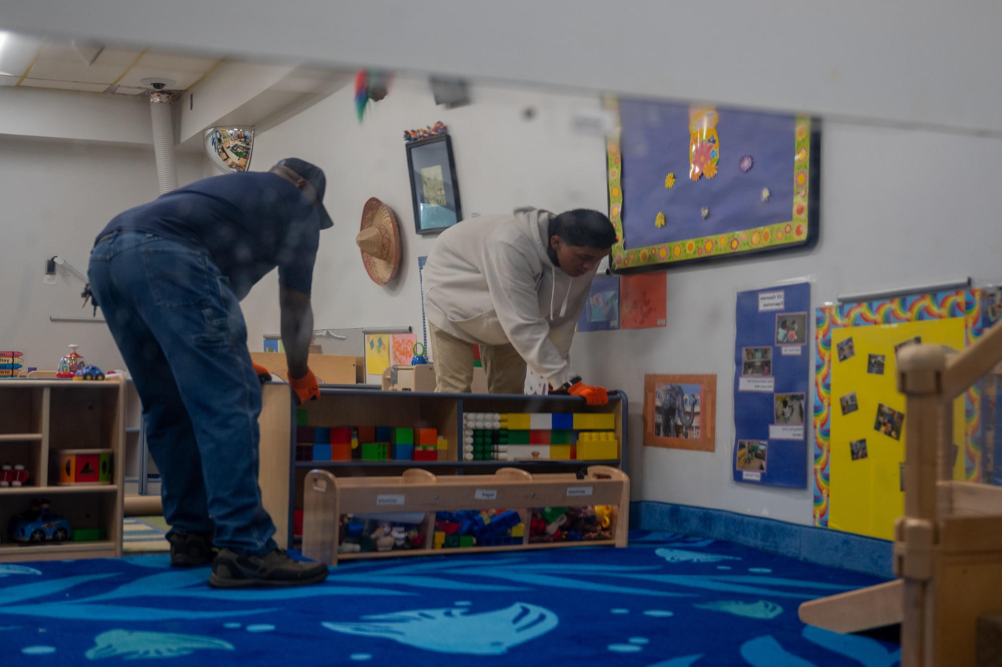 Reflective photo in a mirror of two volunteers in civilian clothing moving a piece of furniture.