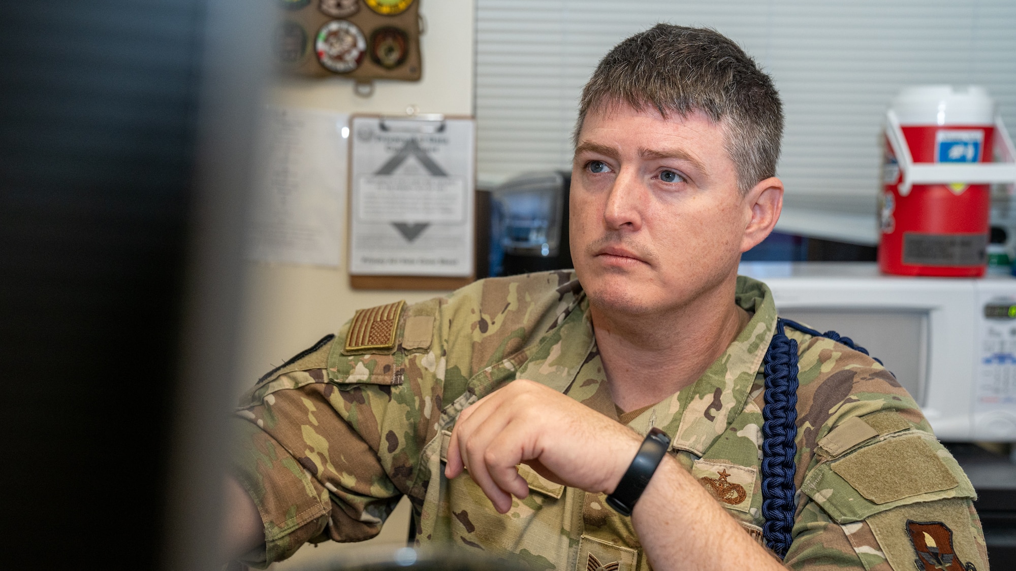 A man in military uniform with a blue rope on his left shoulder that signifies he is a military training leader sits in front of a computer screen.
