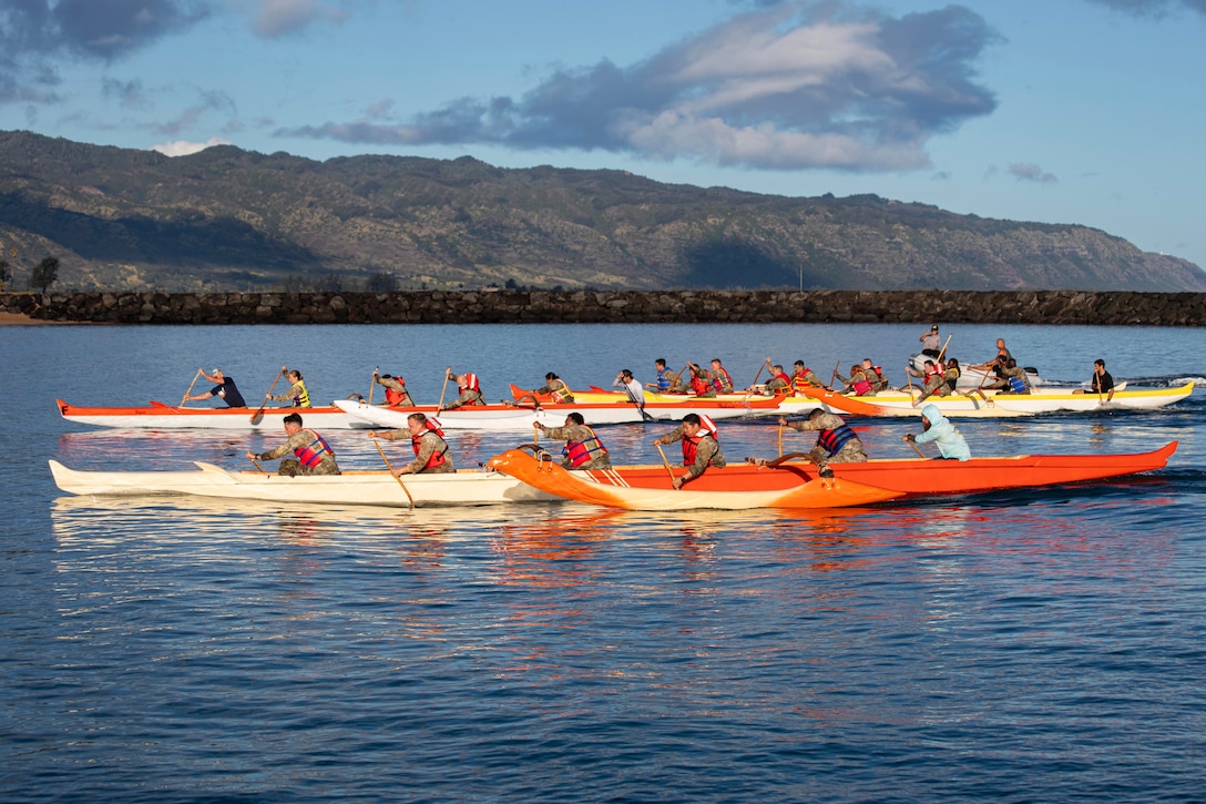 Soldiers in orange and white canoes paddle on the ocean with mountains in the foreground.