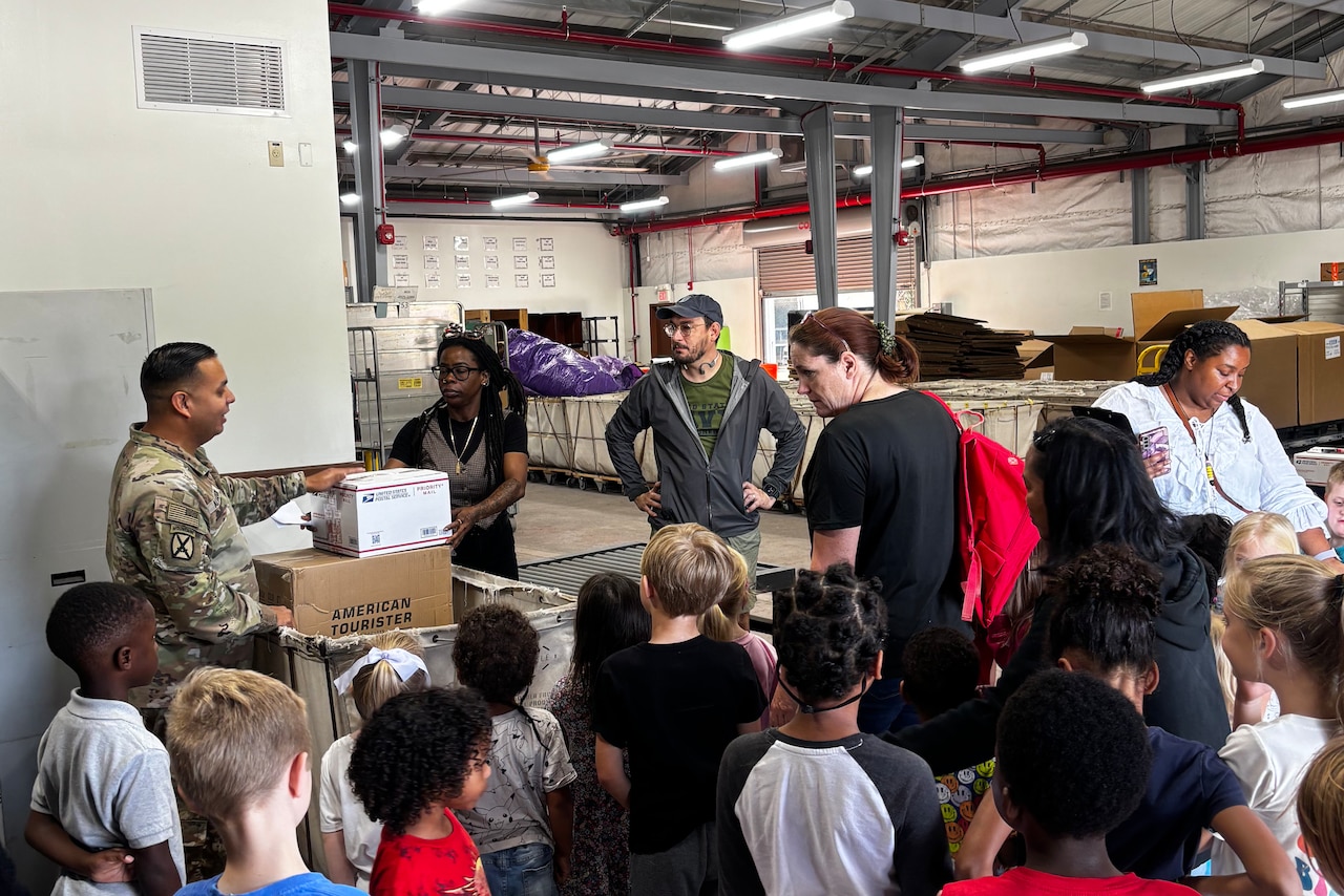 Dozens of students stand in front of adults in a warehouse.