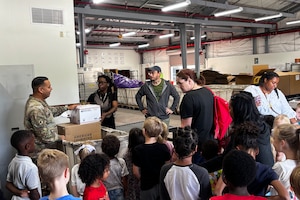 Dozens of students stand in front of adults in a warehouse.