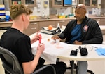 Womack Certified Occupational Therapist Assistant, Reginald Jones (R) works with a Soldier during a therapy session on April 8, 2025. The exercise involves using a 'pickle picker' tool to pick up beads and drop them into a cup.
