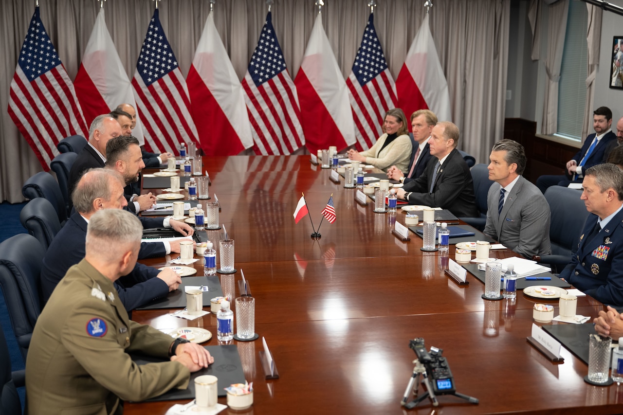 A group of people sit a conference table with flags in the background.