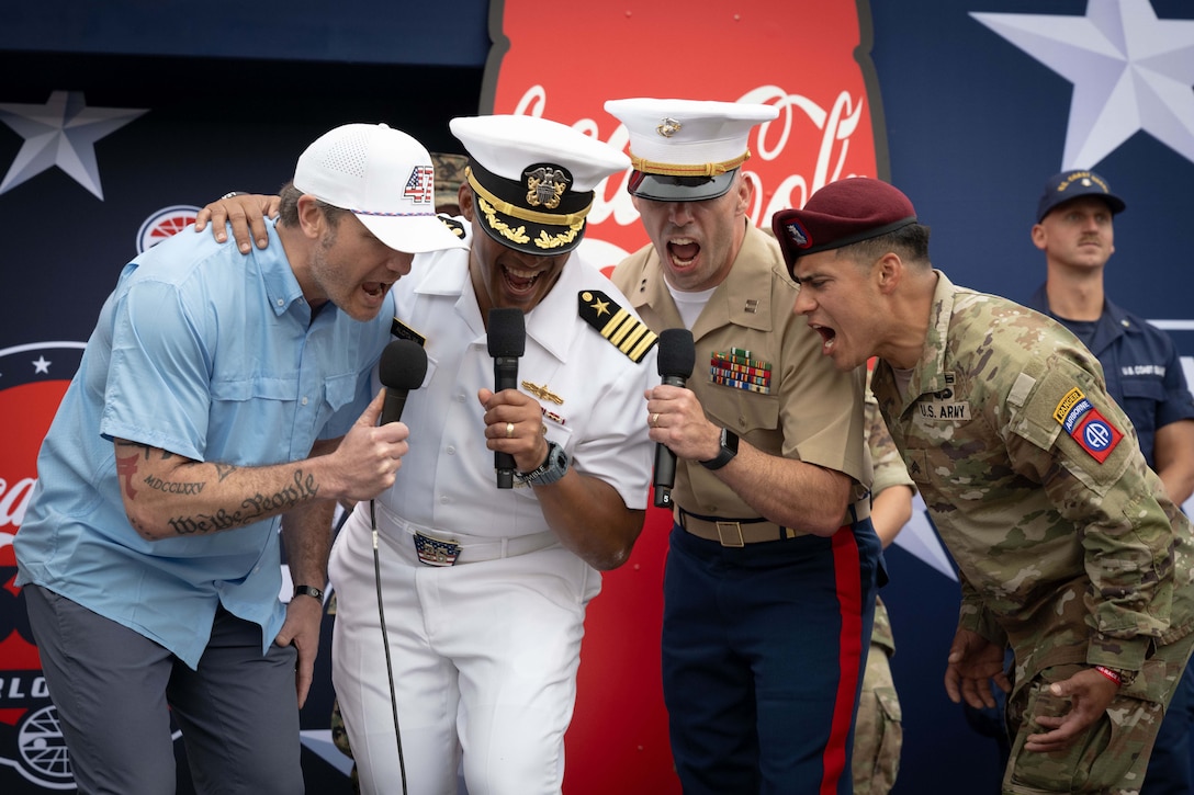 Secretary Pete Hegseth and three service members in uniform yell into microphones in front of a red, white and blue display outdoors.